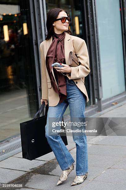 Guest wears brown satin blouse, blue jeans, beige blazer, beige Bottega Veneta clutch bag, snake skin print leather boots, outside Coperni, during...