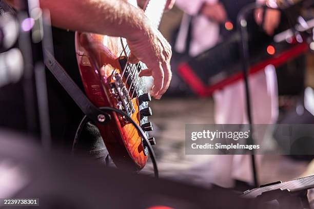 a musician's hand playing a bright maroon-red electric bass guitar, with the figure of another band member dressed in white on stage blurred in the background. - electric guitar stock pictures, royalty-free photos & images