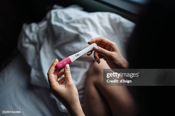 over the shoulder view of an asian woman sitting on bed holding a positive pregnancy test at home. expectant mother anticipating a baby. family concept. life events, motherhood and parenthood. fertility and successful ivf treatment - zwangerschapstest stockfoto's en -beelden