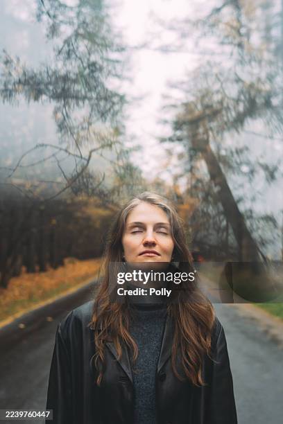 a woman with closed eyes stands on a road, a blurry, moody forest stretching behind her. - midlife crisis stock pictures, royalty-free photos & images