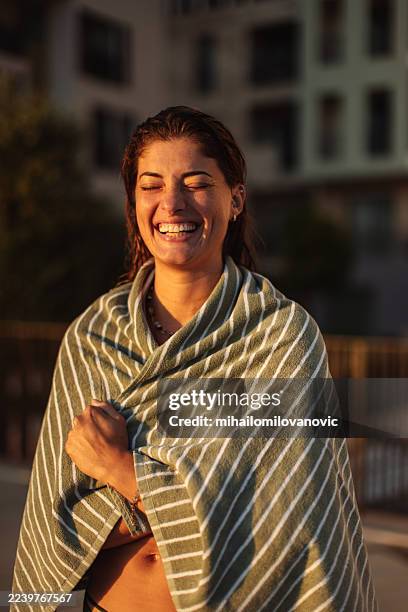 mujer alegre envuelta en toalla a rayas disfrutando del resplandor del atardecer en la azotea de la ciudad al aire libre - envuelto en una toalla fotografías e imágenes de stock