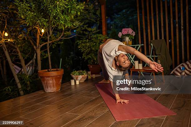 woman practicing yoga outdoors on wooden deck surrounded by plants and lights - homeostasis stock pictures, royalty-free photos & images
