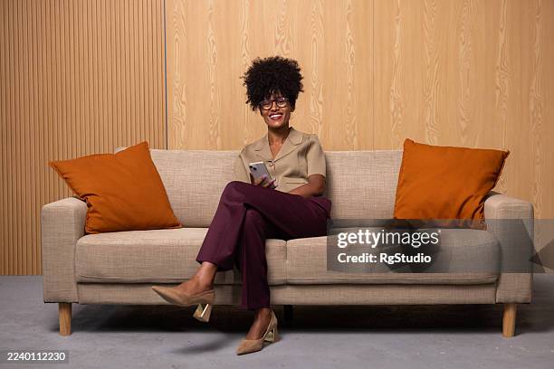 black woman sitting on couch using phone smiling - wide shot stock pictures, royalty-free photos & images