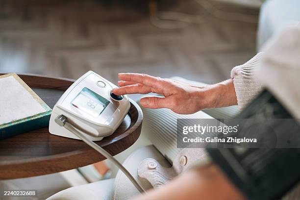 middle-aged woman measuring blood pressure with tonometer at home, sitting on sofa, healthcare concept - hypertensive stock pictures, royalty-free photos & images