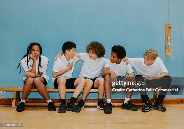 a group of young teenage boys huddle in a sports lesson, deliberately excluding a young lad that sits at the edge of the bench by himself. - stereotypical stock pictures, royalty-free photos & images