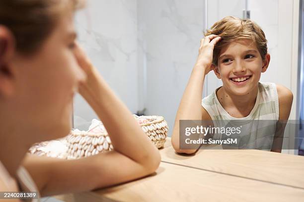 lächelnder junger junge, der mit der hand auf dem kopf in hellem interieur in den badezimmerspiegel schaut - kind vor der pubertät stock-fotos und bilder