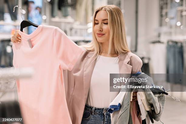 young fashionable shopaholic woman trying on and buying clothes in shopping mall store. consumerism and retail therapy concept in bright store interior. - accro du shopping photos et images de collection