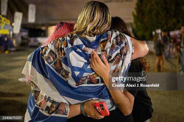 Woman draped in an Israeli flag with hostage faces on it hugs two people as tribute videos as shown while waiting for a live stream of the hostage...
