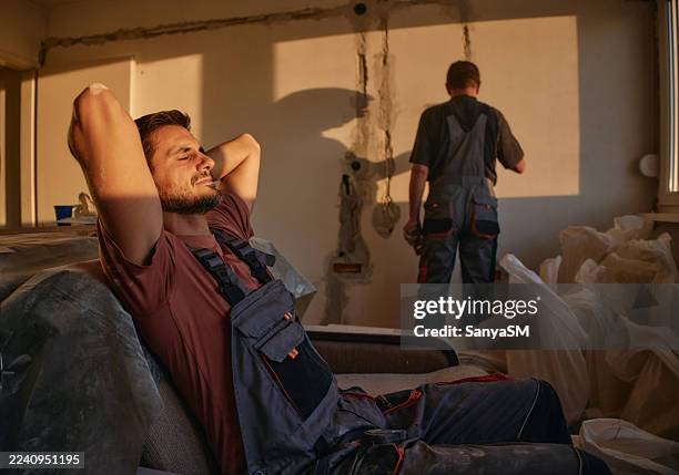entspannter männlicher arbeiter ruht sich auf der couch aus, während kollege bei warmem licht an der renovierung arbeitet - warmes licht stock-fotos und bilder