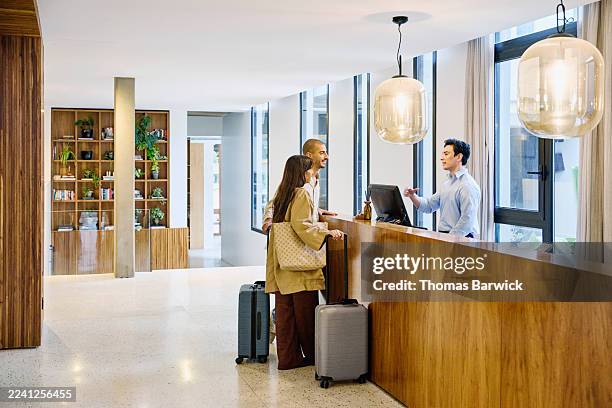 wide shot tourist couple checking in at hotel reception desk - recepção de hotel imagens e fotografias de stock