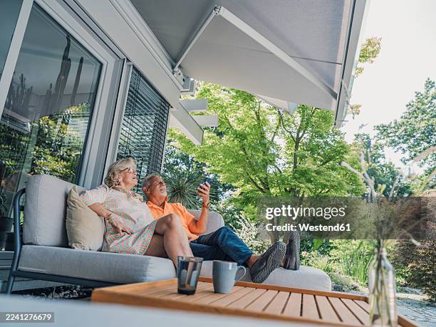 senior couple relaxing on a patio lounge with smartphone and sun protection awning - awning stock pictures, royalty-free photos & images
