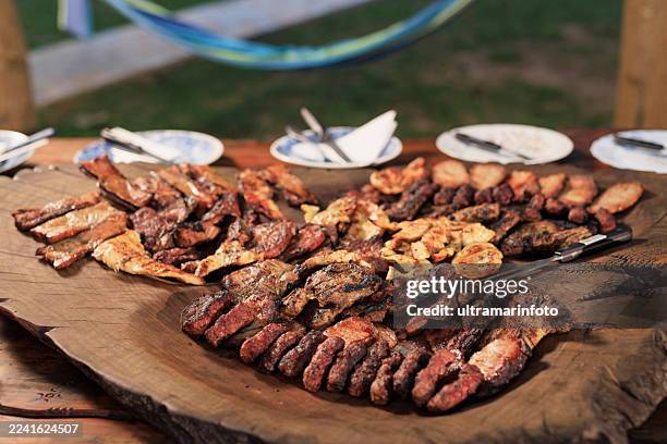 grilled meat served on a wooden board ready for dinner. in the background, a picnic table set up with plates and cutlery - barbecue pork stock pictures, royalty-free photos & images