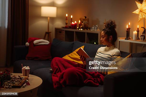 cozy christmas night: woman reading book on sofa with warm lights, blankets and candles - hygge stockfoto's en -beelden