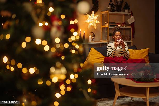 cozy christmas evening: a woman relaxing on a sofa with a mug amid warm lights and festive decor - hygge stockfoto's en -beelden