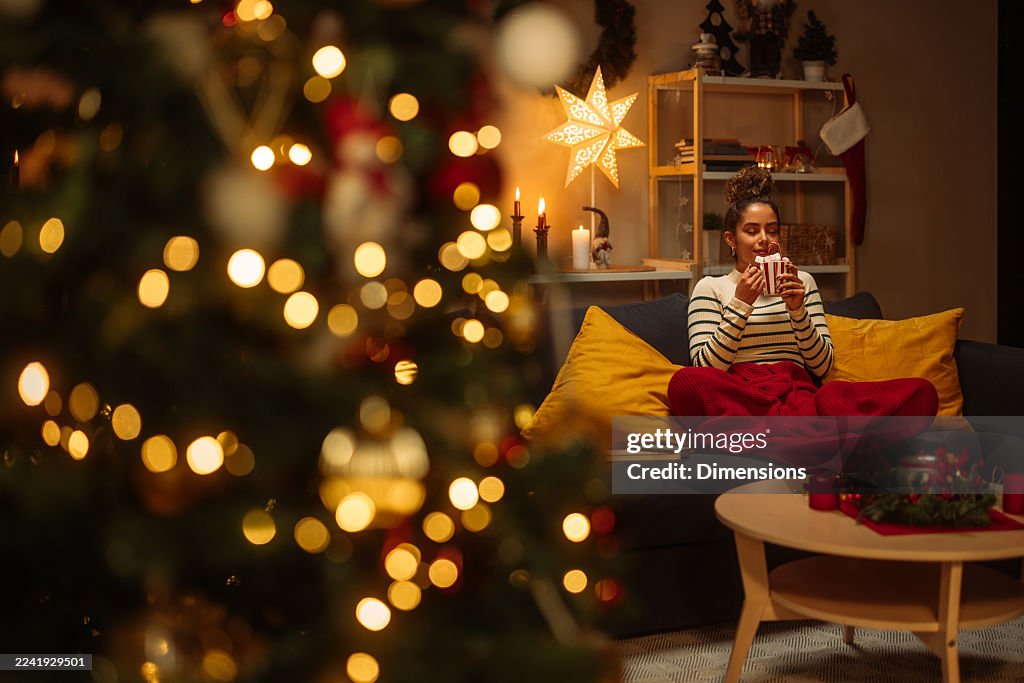 Cozy Christmas evening: a woman relaxing on a sofa with a mug amid warm lights and festive decor