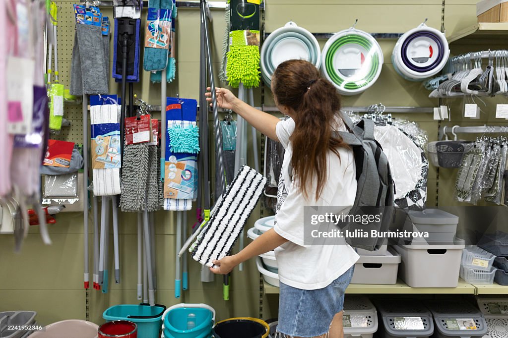 Woman shopping for cleaning supplies in a store, reaching for a mop handle with backpack on her back