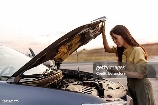 frustrated asian woman stranded beside her broken car, calling for roadside assistance on smartphone - stranded stock pictures, royalty-free photos & images