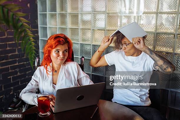 bright red-haired woman works on laptop while friend playfuly tilts notebook over head in casual cafe scene - teasing stock pictures, royalty-free photos & images