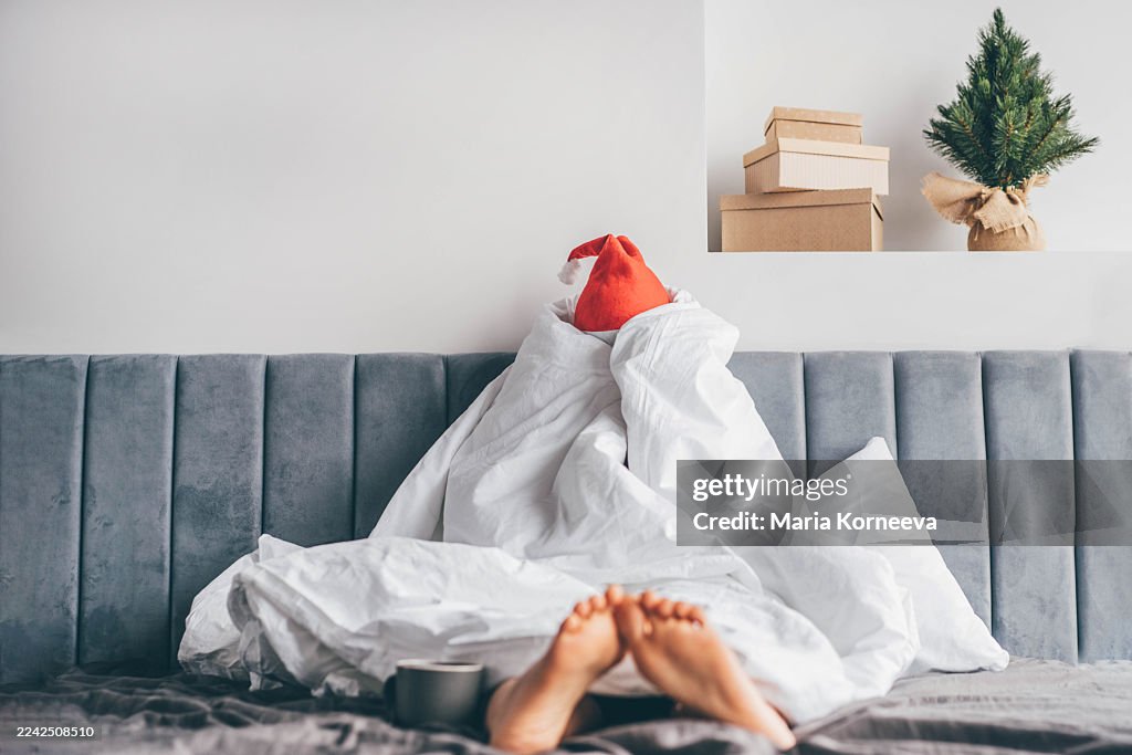 A woman is sitting up in bed, wrapped completely in a white duvet and wearing a Santa hat with a moody, bored expression.