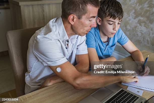 father with diabetes helping his teenage son to do homework or school project using laptop. father is wearing sensor on the back of his upper arm for continuous glucose monitoring. - type 1 diabetes stock pictures, royalty-free photos & images