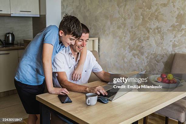 teenage boy hugging his father with diabetes while working on laptop for homework or school project. father is wearing sensor on the back of his upper arm for continuous glucose monitoring. - type 1 diabetes stock pictures, royalty-free photos & images