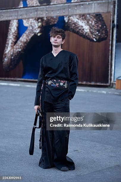 Alexey Sorochin wears a black wrap-style top paired with wide-leg black trousers, accessorised with a floral embroidered sash belt and a black...