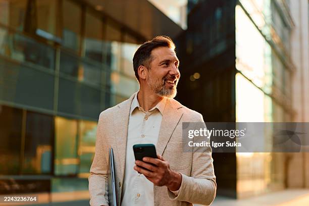 smiling businessman walking in city with smartphone and laptop - golden hour stock pictures, royalty-free photos & images