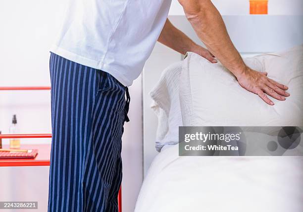 man in striped pajamas arranges pillow while making bed in bright modern bedroom setting - assembling a bed frame stock pictures, royalty-free photos & images