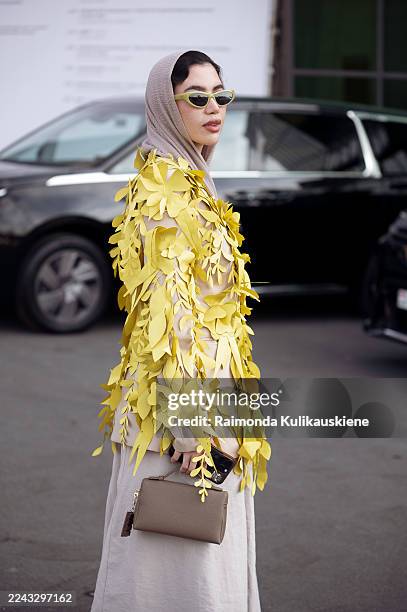 Kiana Eva wears a beige maxi skirt and matching top layered under a striking yellow coat covered in 3D leaf appliqués, styled with a taupe headscarf,...