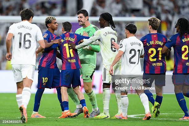 Lamine Yamal of FC Barcelona is held back by Marc Casado of FC Barcelona as players of Real Madrid Thibaut Courtois and Eduardo Camavinga of Real...