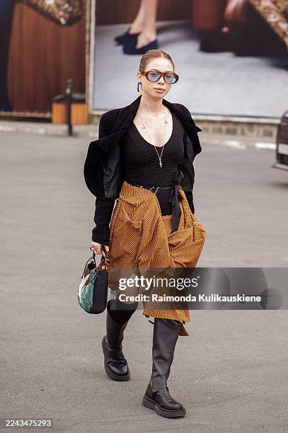 Guest wears a black textured jacket over a brown and black check wrap skirt with a slit, styled with a mesh top, black knee-high boots, oversized...