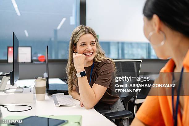 woman smiling, laughing and talking with a colleague at office desk in a modern workplace setting - bedrijfscultuur stockfoto's en -beelden