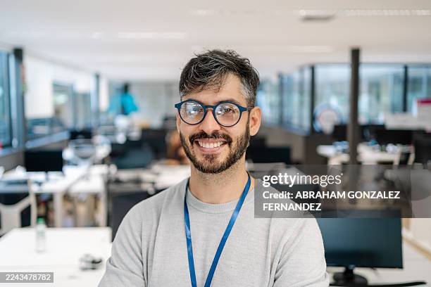 smiling male professional wearing glasses and id badge, standing in a modern open plan office - headshot stock pictures, royalty-free photos & images