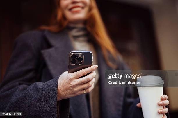 frau im mantel benutzt smartphone im freien mit kaffeetasse auf einer belebten städtischen straße - halbnahe-einstellung stock-fotos und bilder