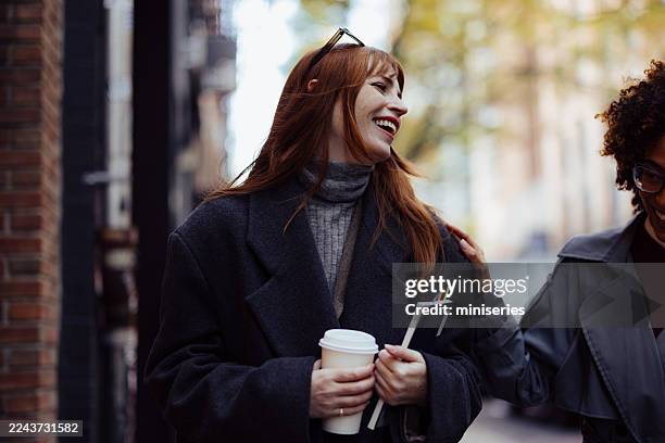 two friends laughing on a busy street with coffee cup and notebook in cozy outerwear - long coat stock pictures, royalty-free photos & images