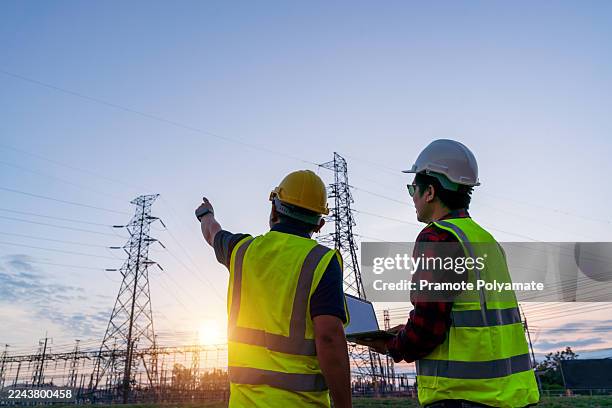 two electrical engineers discuss energy planning using a laptop, pointing at high-voltage transmission towers and the sun flare, technology-driven teamwork and grid modernization at power substation. - smart grid foto e immagini stock