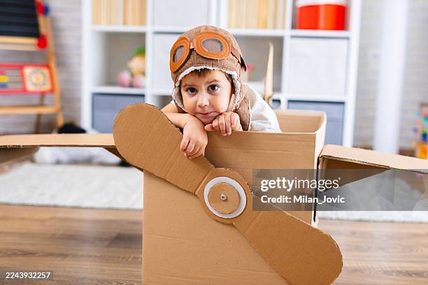 cute boy dressed as a pilot pretends to fly in a cardboard box airplane, showing creativity and childhood imagination. - aviation hat stock pictures, royalty-free photos & images