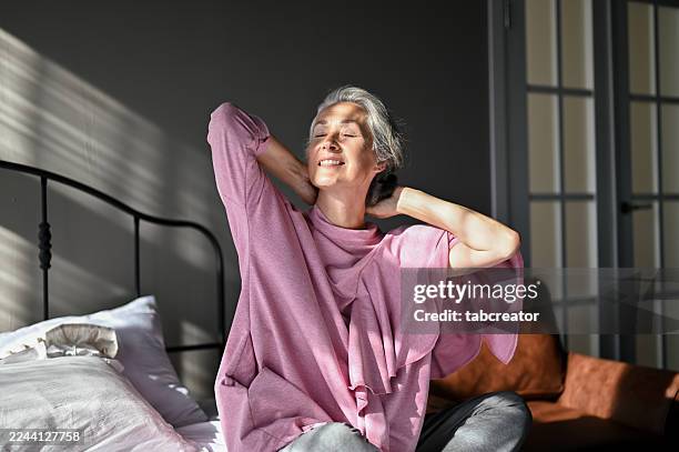 mature adult woman stretching on bed in sunny bedroom - atmospheric mood stock pictures, royalty-free photos & images