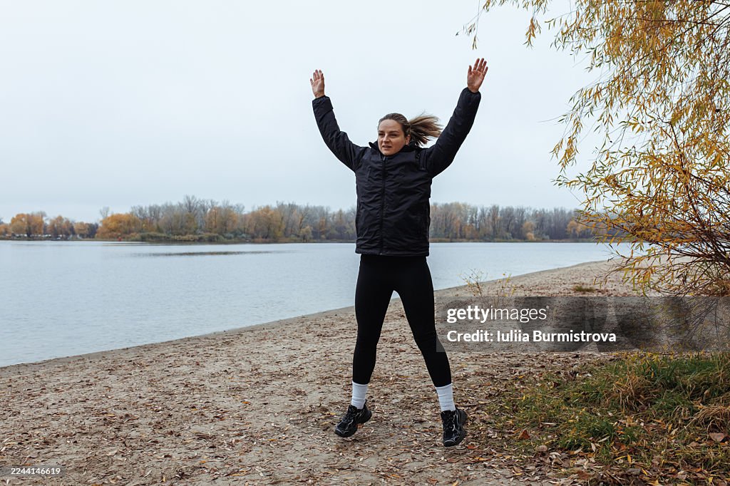Woman jumping with arms raised on autumn lakeside path, expressing energy and joy