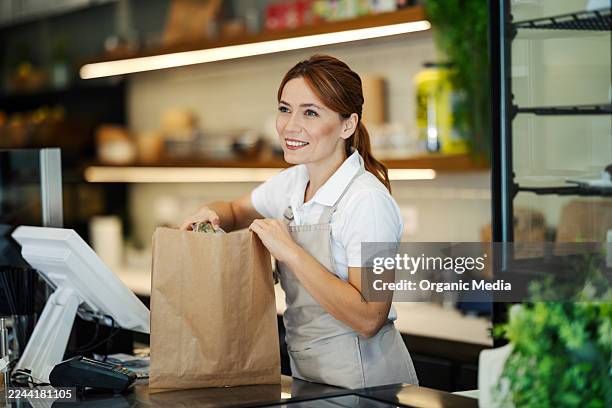 woman cashier packing groceries in paper bag at store checkout - frontline worker stock pictures, royalty-free photos & images