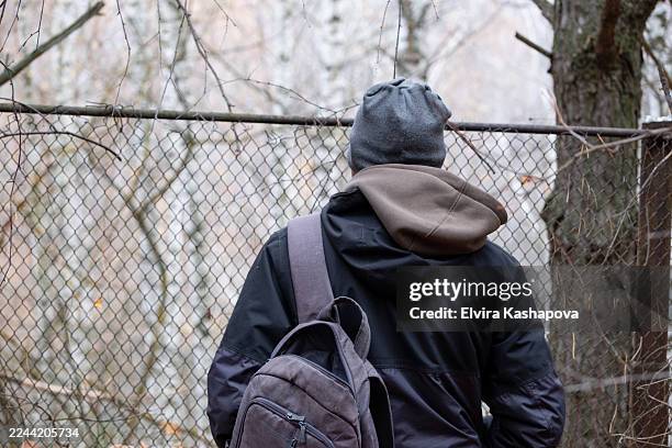 a man wearing a jacket and carrying a backpack stands in front of a border crossing fence in the forest. - emigración e inmigración fotografías e imágenes de stock