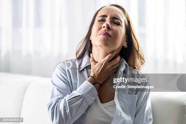 a woman is sitting on a sofa, her eyes closed and her expression frowning, holding her hand on her throat. she seems to be experiencing pain or a scratchy feeling in her throat, which indicates a medical condition such as a sore throat, dry cough. - schilddrüsenüberfunktion stock-fotos und bilder