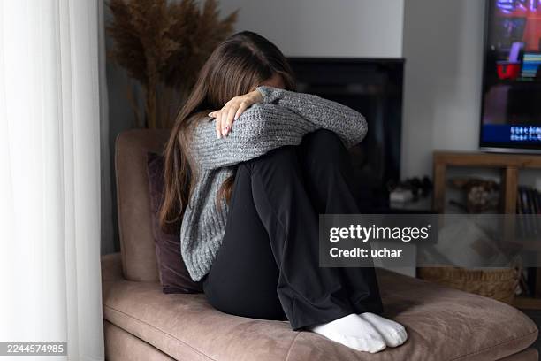 young woman feeling sad and anxious, sitting alone on a couch at home - shy stock pictures, royalty-free photos & images