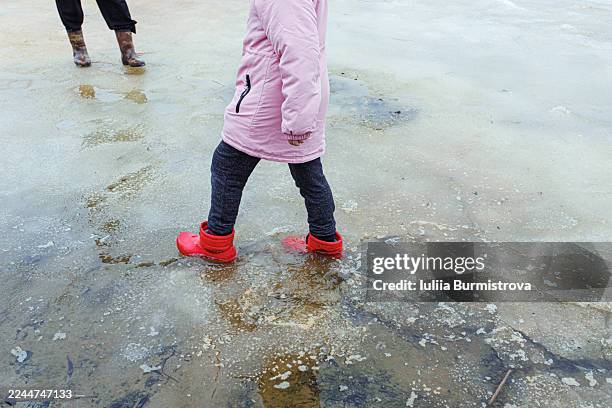 child in pink coat and red boots walking on melted ice outdoors with adult legs nearby - long coat stock pictures, royalty-free photos & images