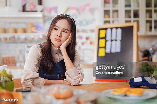 hardworking bakery owner focuses on her laptop inside the cake shop, her face showing stress and determination as she manages orders and business tasks. nearby, a dedicated male baker and a thoughtful korean barista work diligently reflecting challenge - struggle stock pictures, royalty-free photos & images
