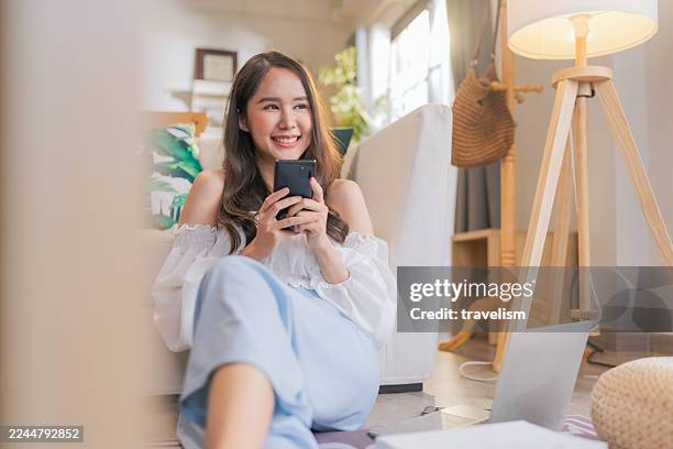 une jeune femme heureuse est assise confortablement sur le sol de la maison, souriante tout en tenant son téléphone portable et en dégustant une boisson rafraîchissante. entourée d’une atmosphère chaleureuse et détendue, elle adopte un style de vi - aplatir la courbe photos et images de collection