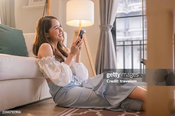 une jeune femme heureuse est assise confortablement sur le sol de la maison, souriante tout en tenant son téléphone portable et en dégustant une boisson rafraîchissante. entourée d’une atmosphère chaleureuse et détendue, elle adopte un style de vi - aplatir la courbe photos et images de collection