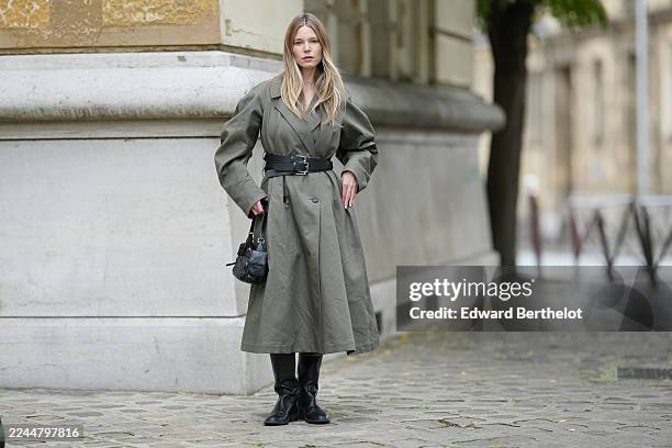 Natalia Verza wears long straight blonde hair, an olive green belted trench long coat with multi belts, black leather knee-high boots by Giuseppe...