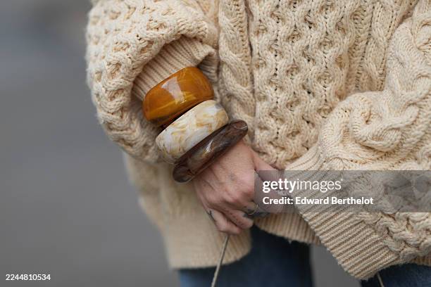 Emy Venturini wears three oversized resin bangles in amber, ivory marbled, and brown tones, styled over the sleeve of a chunky cream cable-knit...