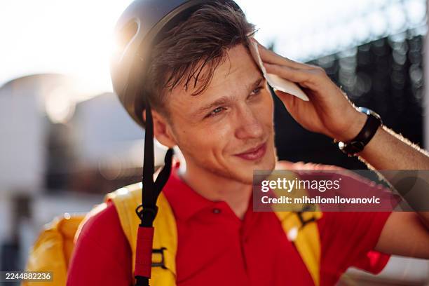 joven con casco, camisa roja y mochila amarilla limpiándose el sudor al aire libre - entrega sin contacto fotografías e imágenes de stock
